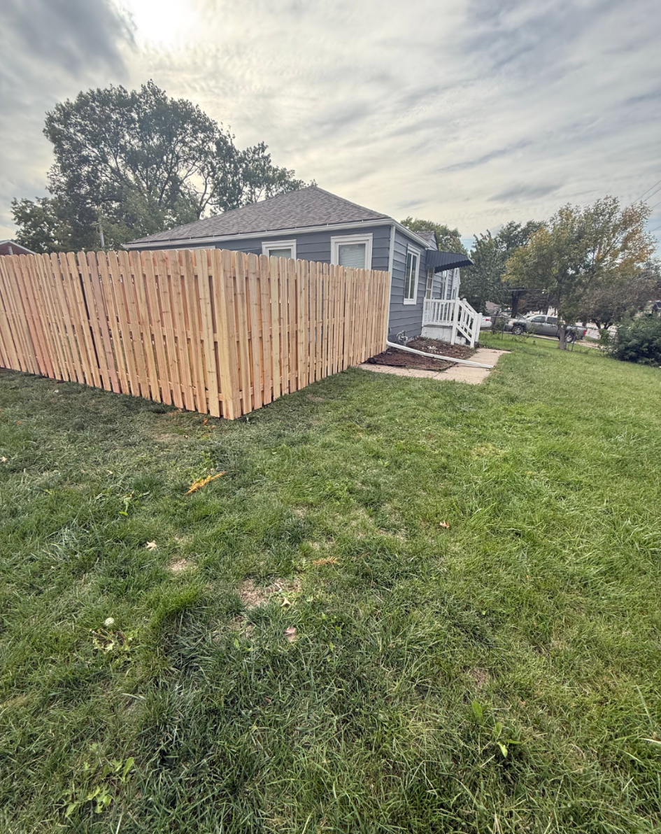 Wooden fence encloses the side yard of a gray house with white trim. Green grass and a cloudy sky.
