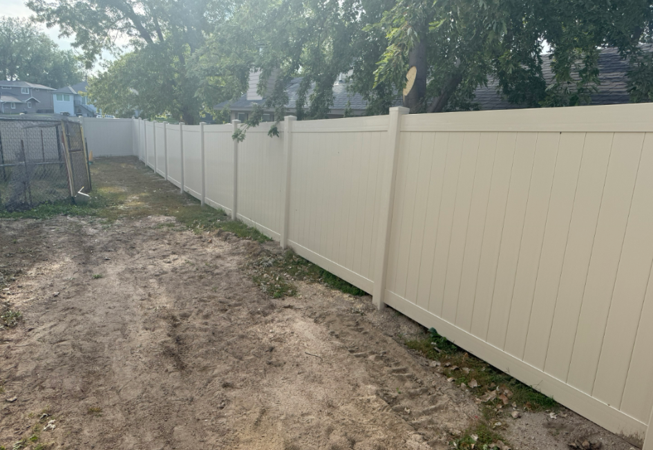 Tan vinyl fence in a yard with dirt and grass, trees in the background.