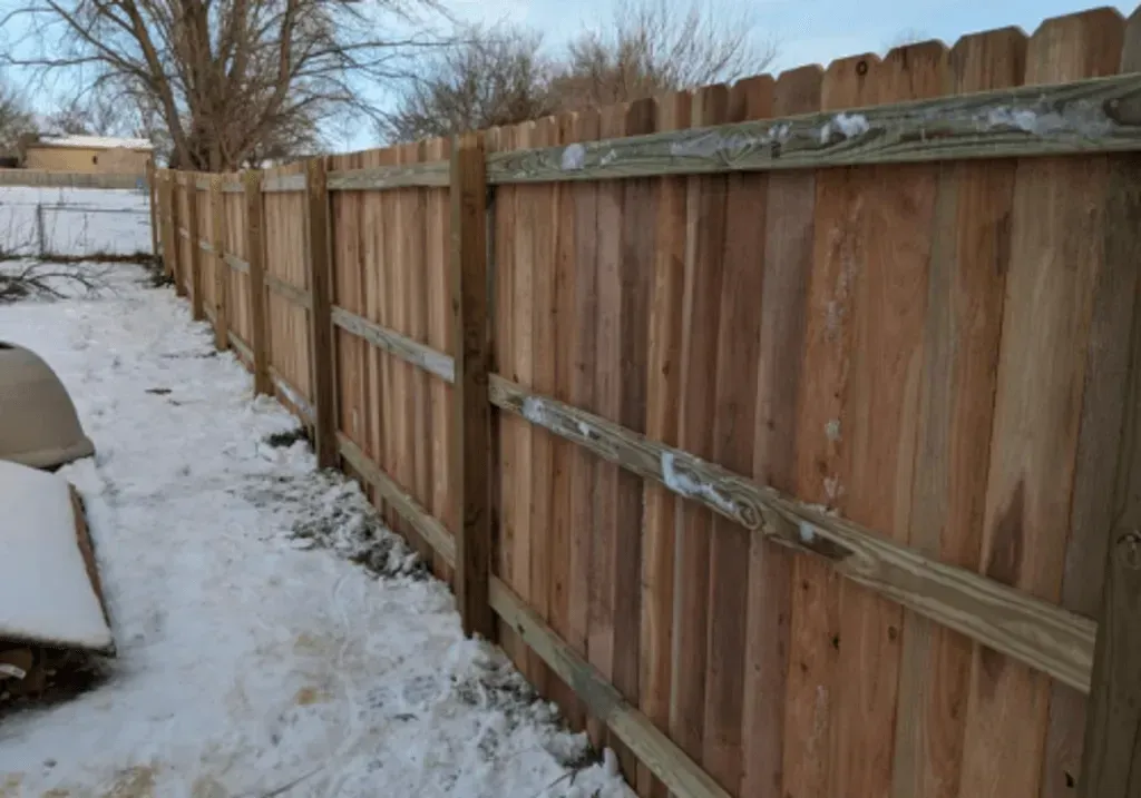 Wooden fence in a snowy yard. Brown planks, horizontal rails, and wooden posts.