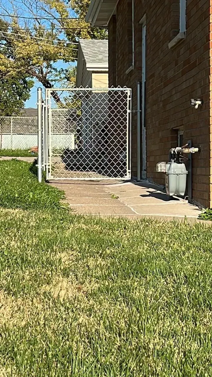 Chain link fence next to a brick building; grassy yard in front. Gas meter on the building.