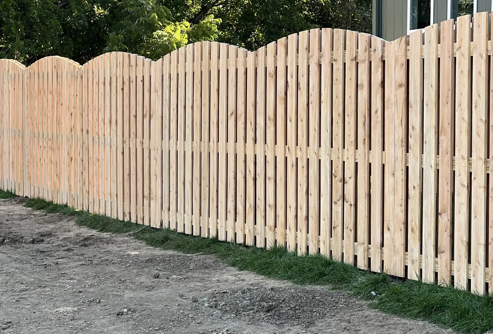 Wooden fence with scalloped top, bordering gravel and grass in a yard.