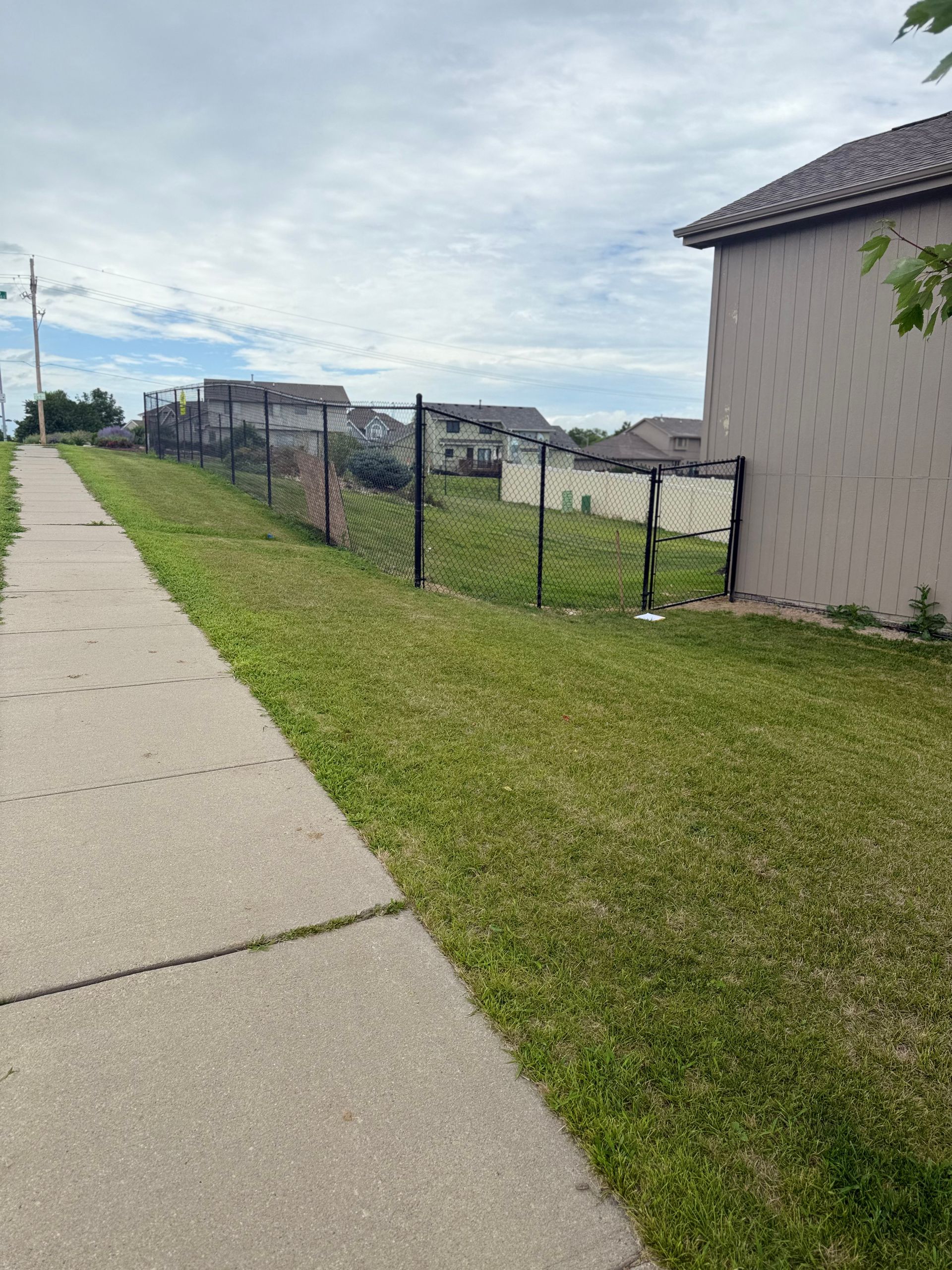 Sidewalk next to a sloped, grassy area with a fence, leading to houses under a cloudy sky.