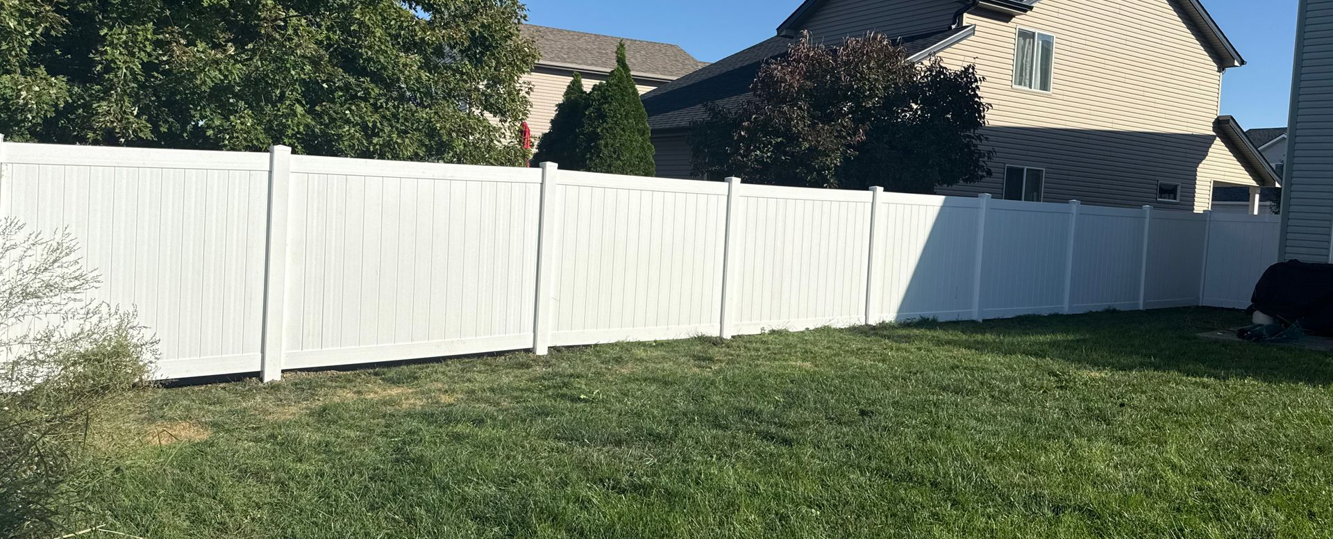 White vinyl fence in a grassy yard, partially obscuring a house and trees under a clear blue sky.