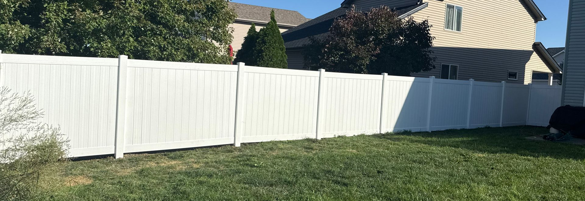 White vinyl fence in a grassy backyard, trees and houses in the background.