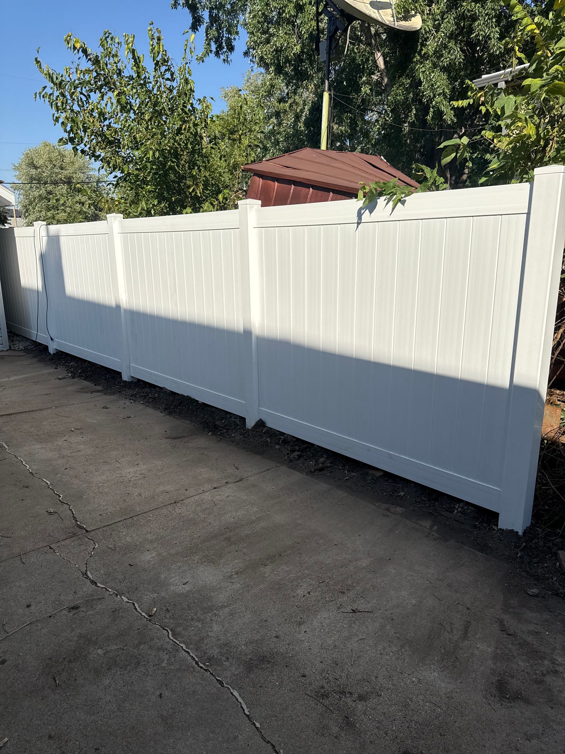 White vinyl fence along a paved driveway with green trees in the background under a blue sky.