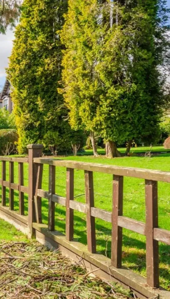 Wooden fence in front of green lawn with trees and a house in the background.