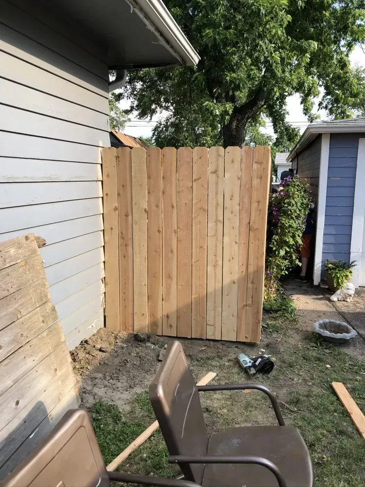 Wooden fence section under construction next to a gray house, with brown chairs and tools nearby.