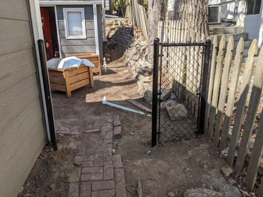A black chain link fence gate opens to a dirt pathway between a house and a wooden fence.