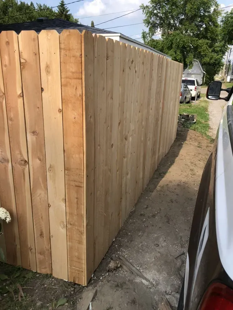 Wooden fence along a paved driveway next to a parked car on a sunny day.