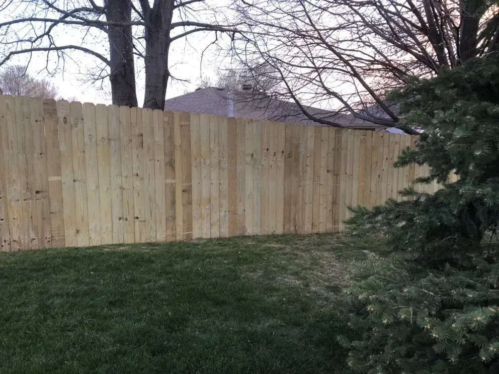 Wooden privacy fence in a grassy yard, with trees and a house in the background.