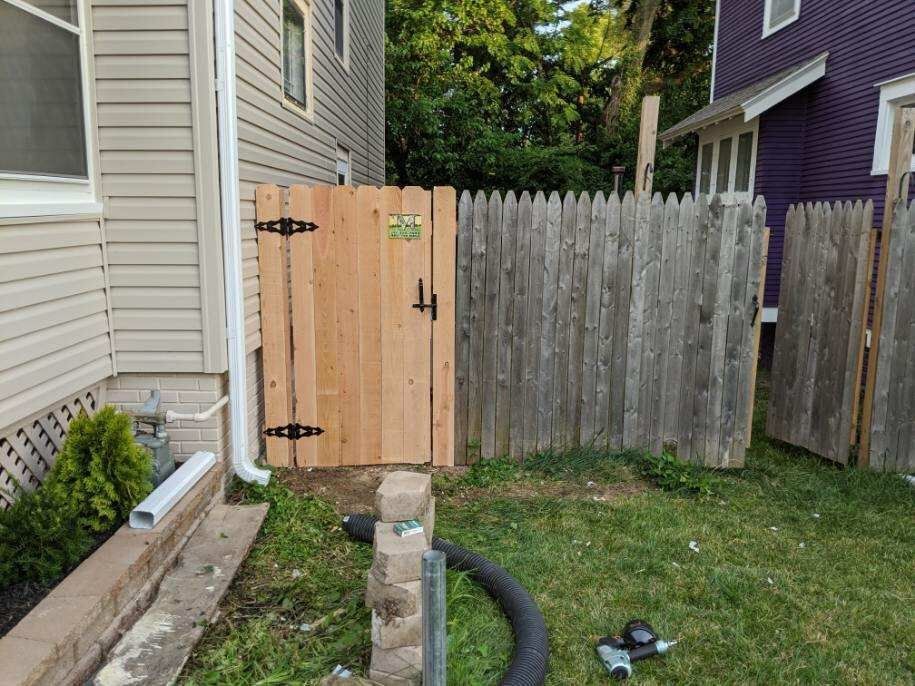 Wooden gate, brown and new, next to a weathered fence. Beige siding on the left. Green grass.