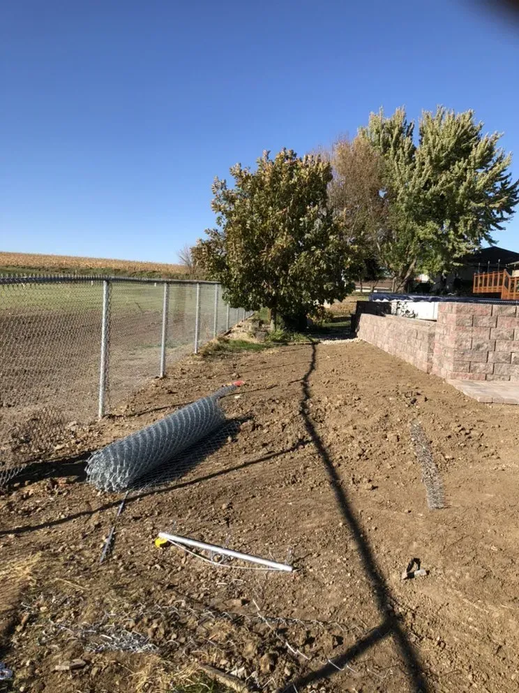 Chain-link fence construction in progress, with rolled wire, posts, and dirt, on a sunny day.