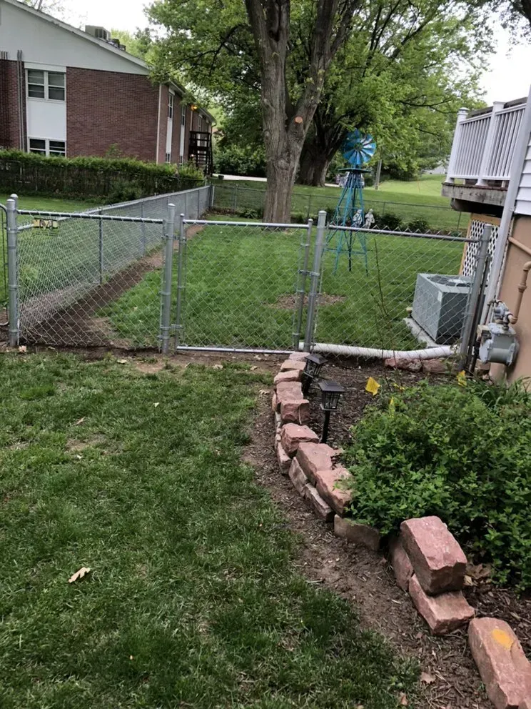 Chain-link fence in backyard with a brick border and a building in the background.