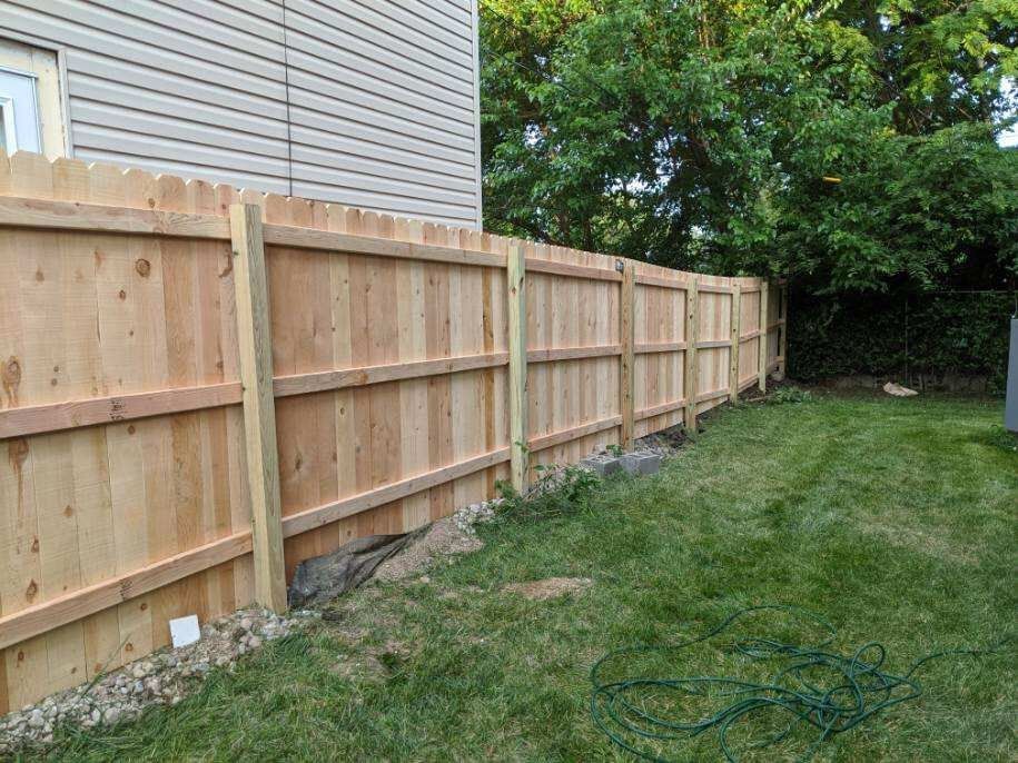 Wooden fence in a grassy yard, beside a beige-sided building. Green trees in the background.