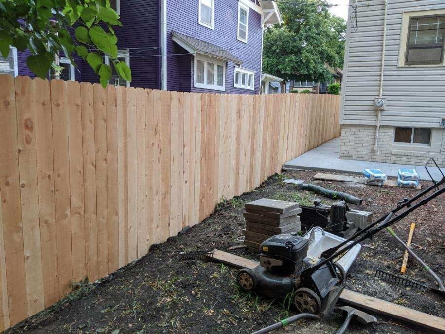 Wooden fence in backyard next to a house with a lawnmower on the ground.
