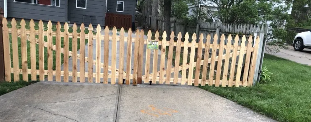 A light brown picket fence curves along a driveway, with a house and green lawn in the background.