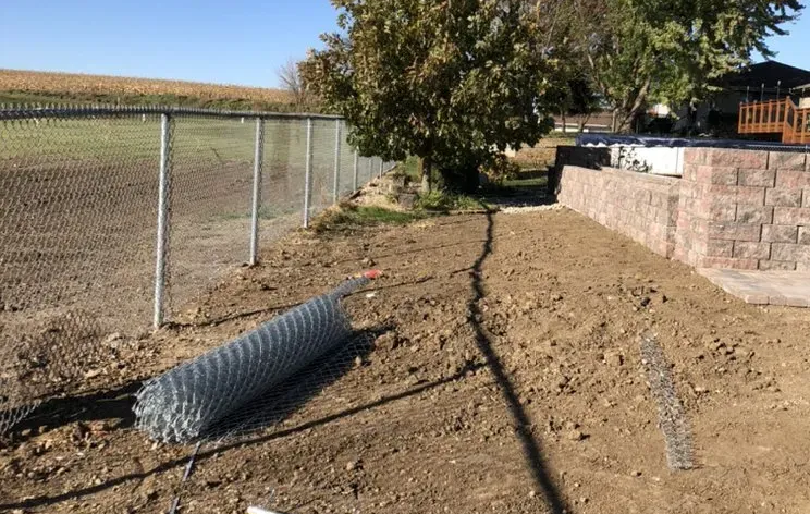 A roll of chain-link fence on the ground near an existing fence and brick wall, dirt path.