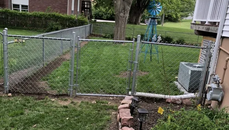 Chain-link fence encloses a grassy backyard with an AC unit, tree, and decorative windmill in the background.