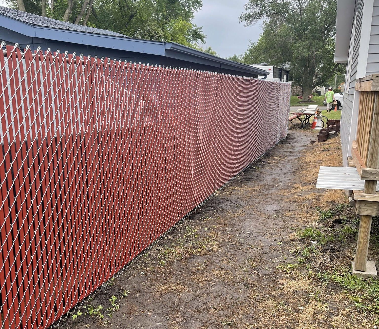 Red chain-link fence running along a building and a muddy path. Overcast sky.