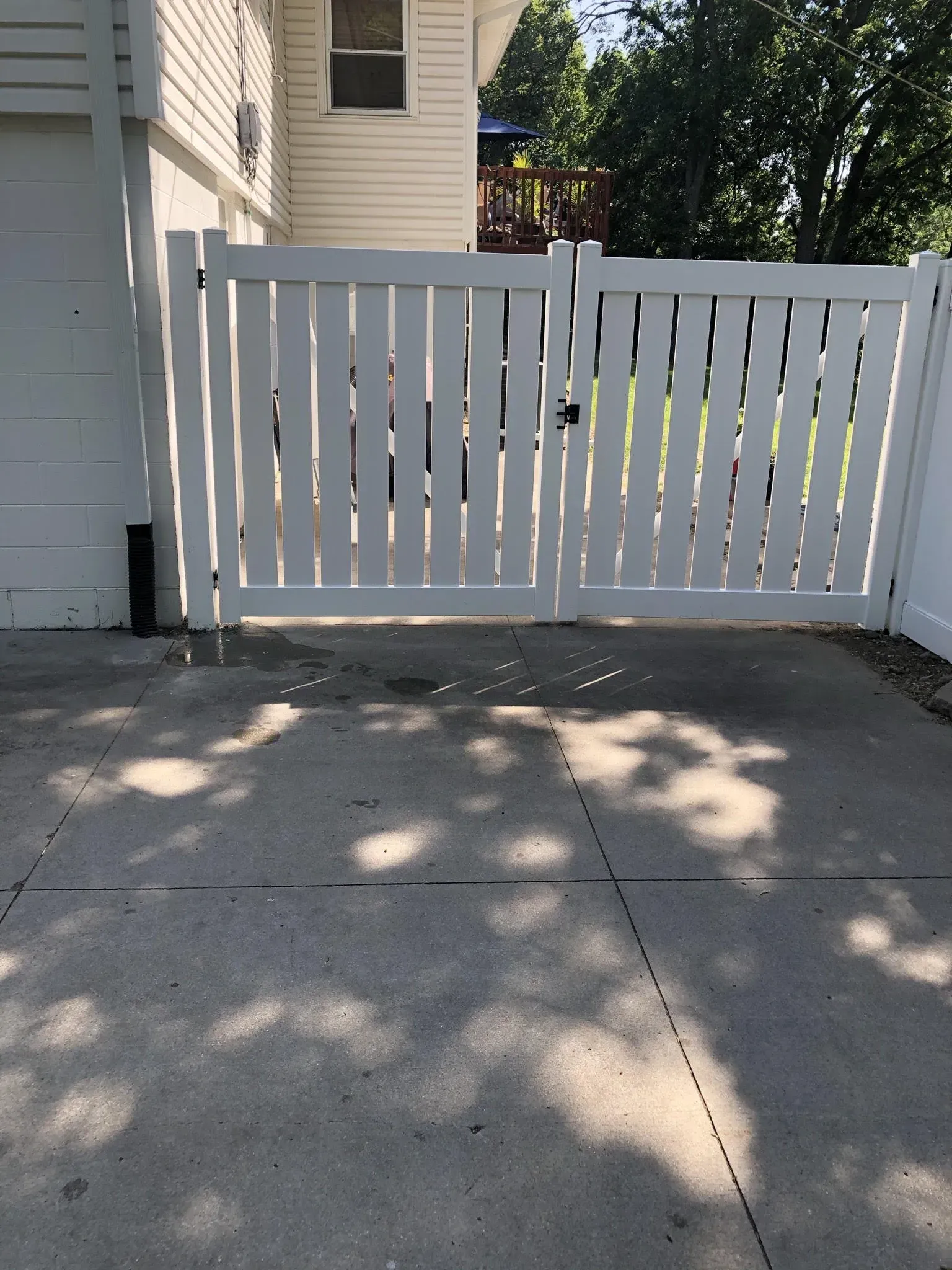 White double gate in a white fence, on a concrete driveway, sunlight overhead.