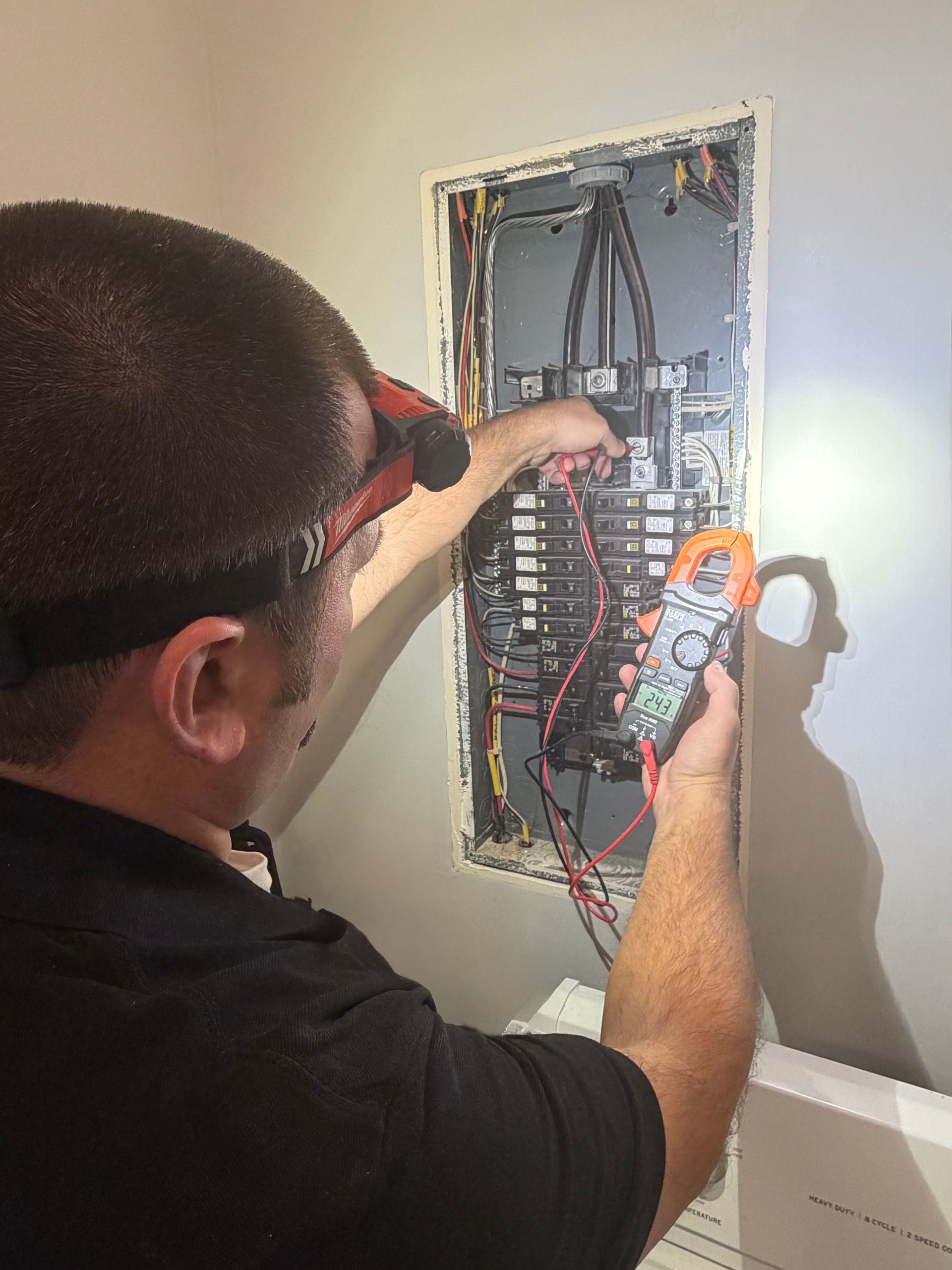 Electrician using a multimeter on an open electrical panel, illuminated by a headlamp.