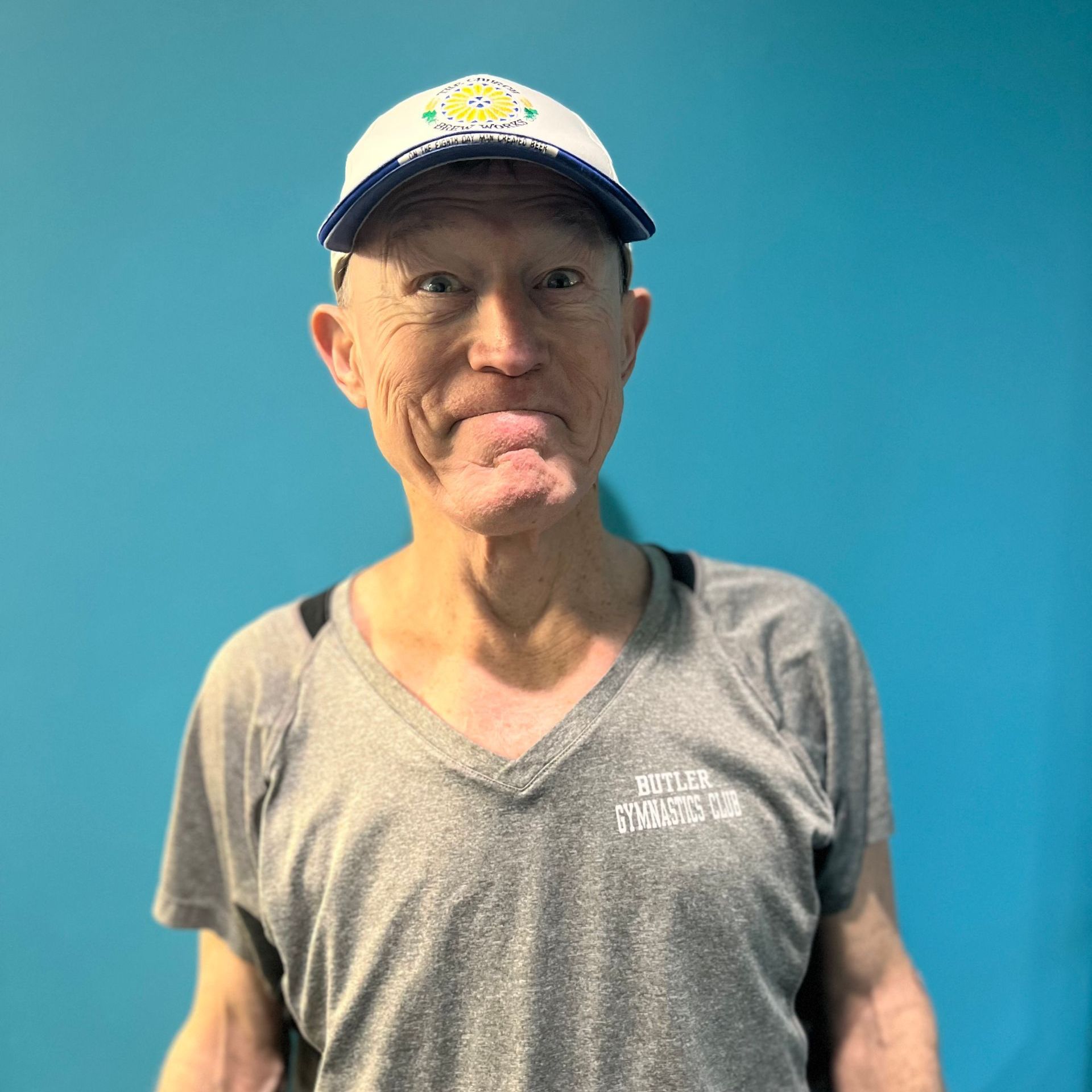 A man wearing a hat and a grey shirt is standing in front of a blue wall.