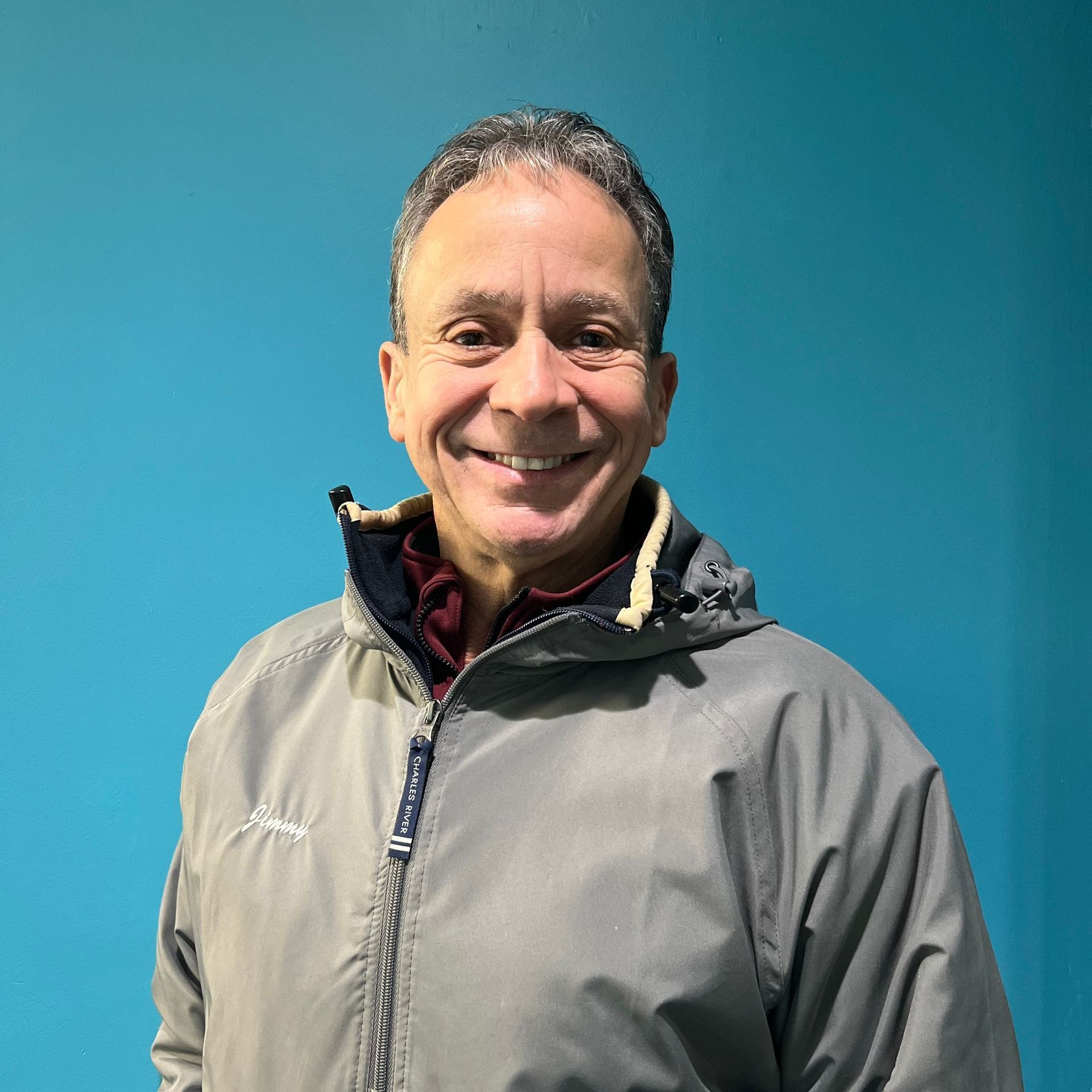 A man in a grey jacket is smiling in front of a blue wall.