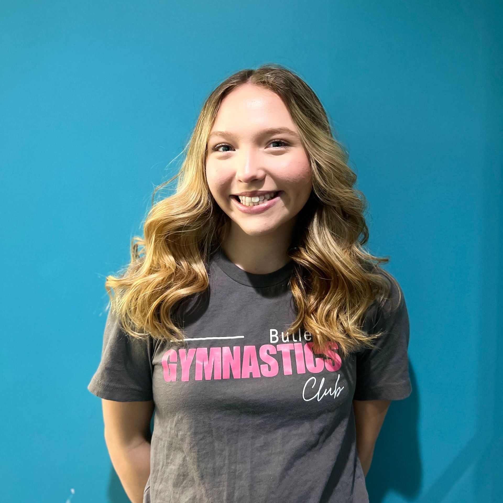 A young woman wearing a gymnastics t-shirt is smiling in front of a blue wall.