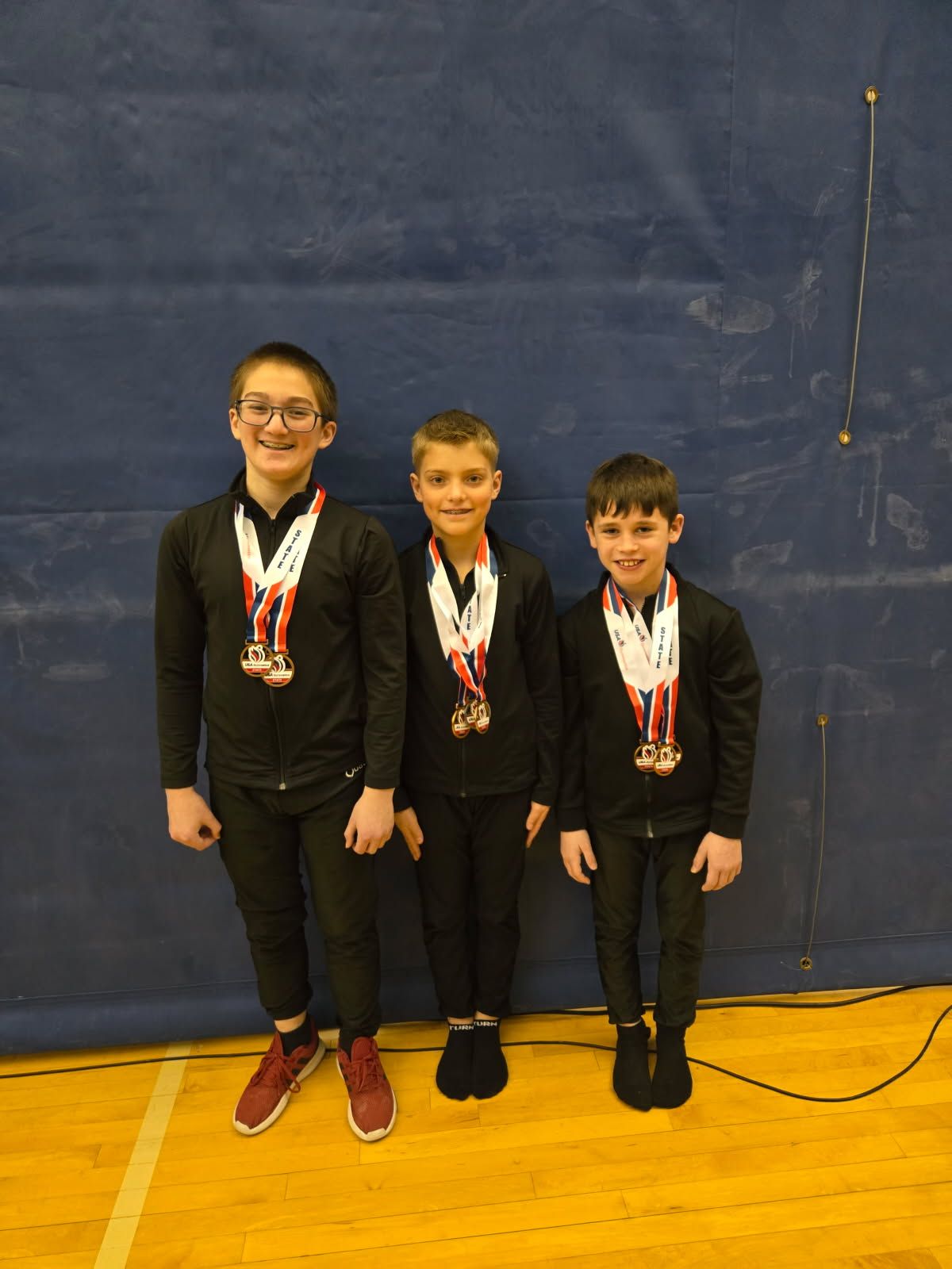 Three young boys wearing medals are standing next to each other on a gym floor.