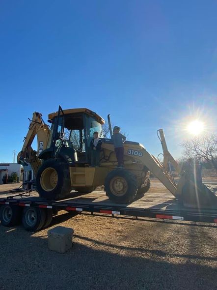 Yellow backhoe on a trailer under a bright blue sky; two people in the cab.