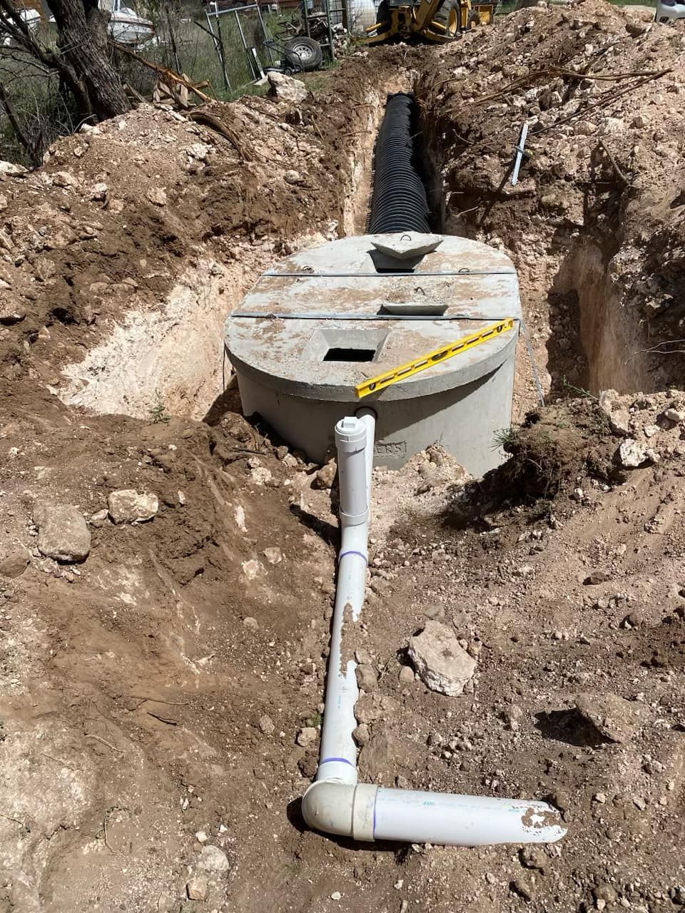 A concrete septic tank installed in a dirt trench, with pipes and construction tools visible.