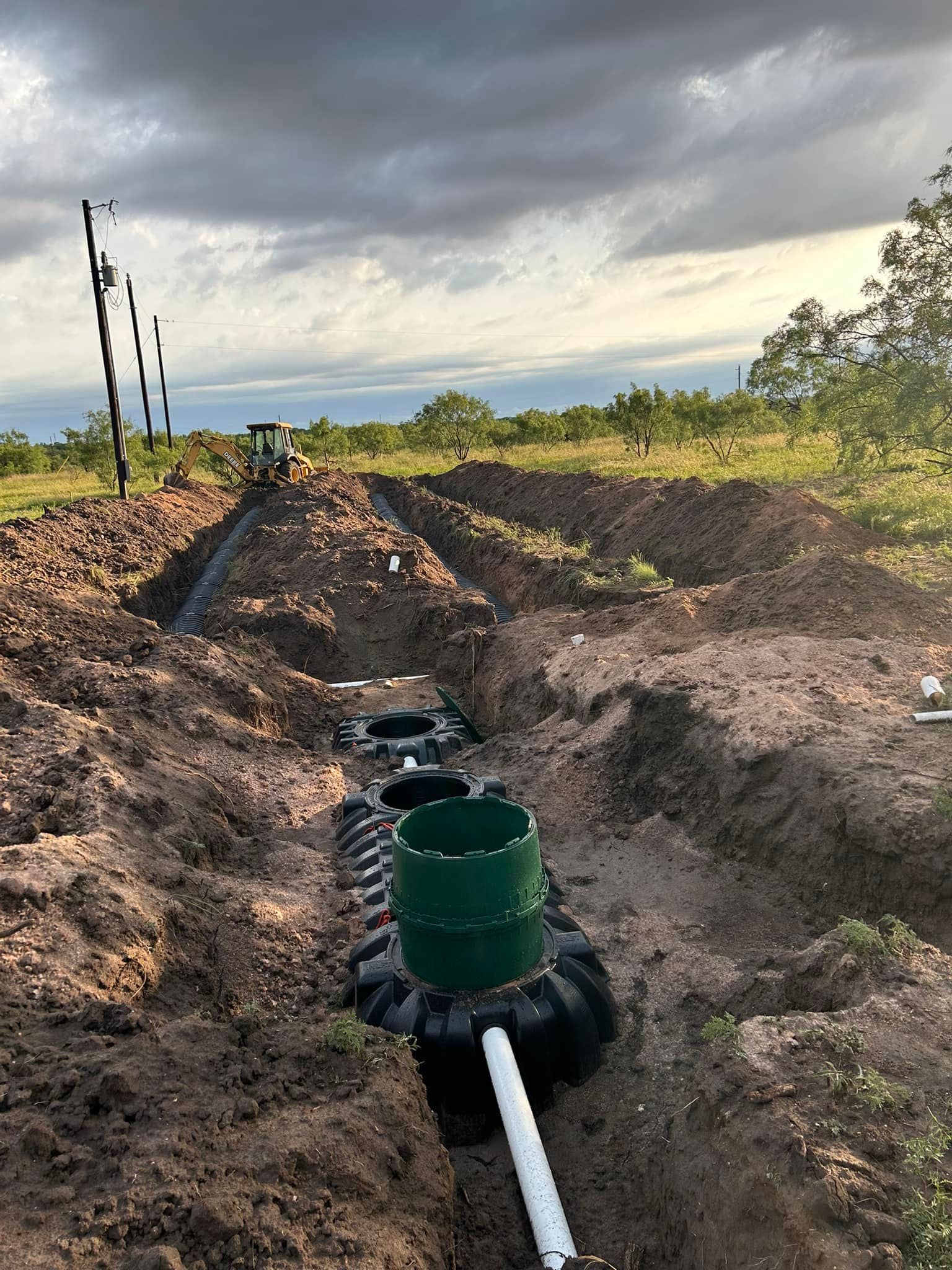 A septic system installation: green tank in a trench, with dirt and equipment in a field. Cloudy sky.