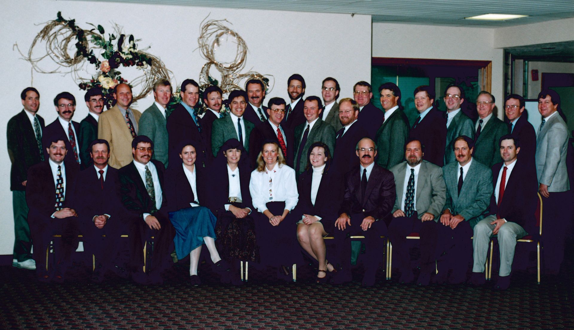 A large group of people in business formal attire posing for a formal portrait in a conference room with decor on the wall.