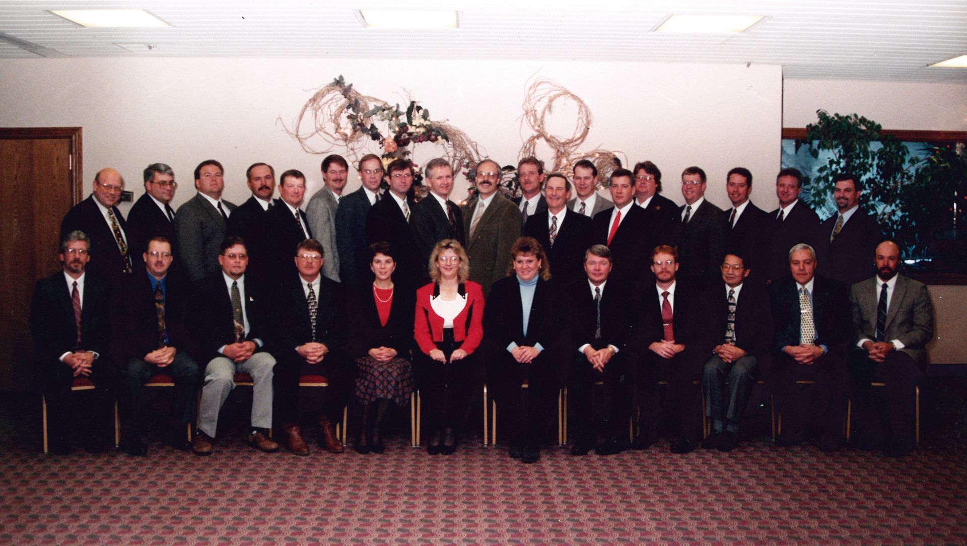 A group of people in business attire posing for a formal photo in a room with a patterned wall sculpture.