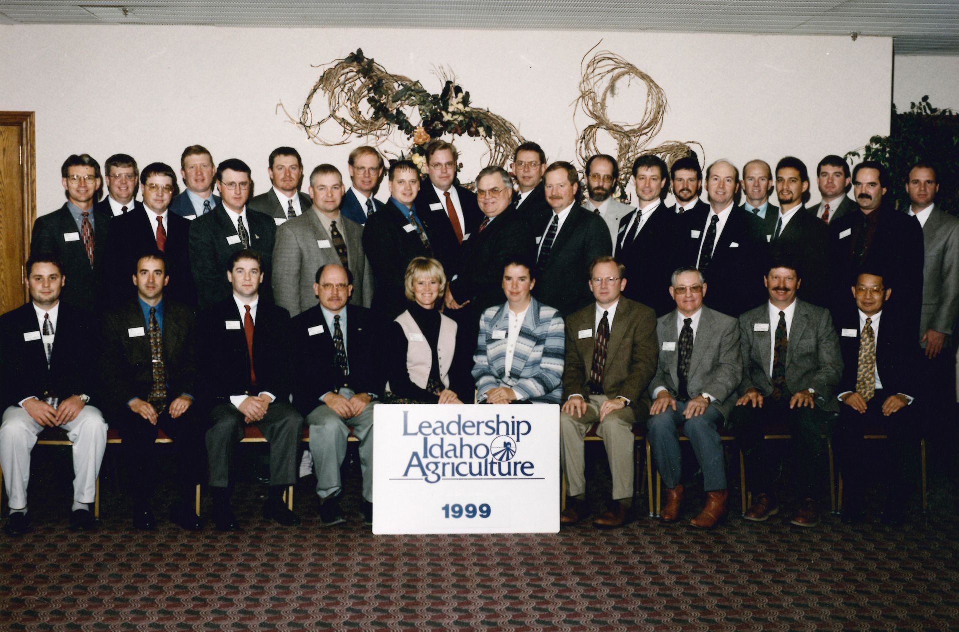 A group of people pose for a photo in front of a Leadership Idaho Agriculture 1999 sign at an indoor gathering.