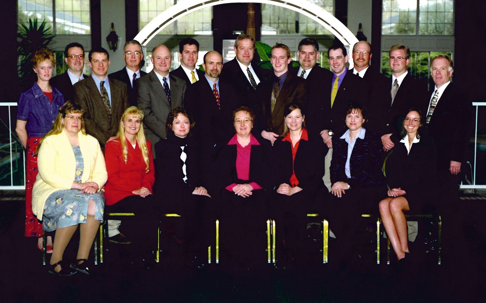 A group of professional individuals posing for a formal portrait in an elegant indoor venue with a large arched window.