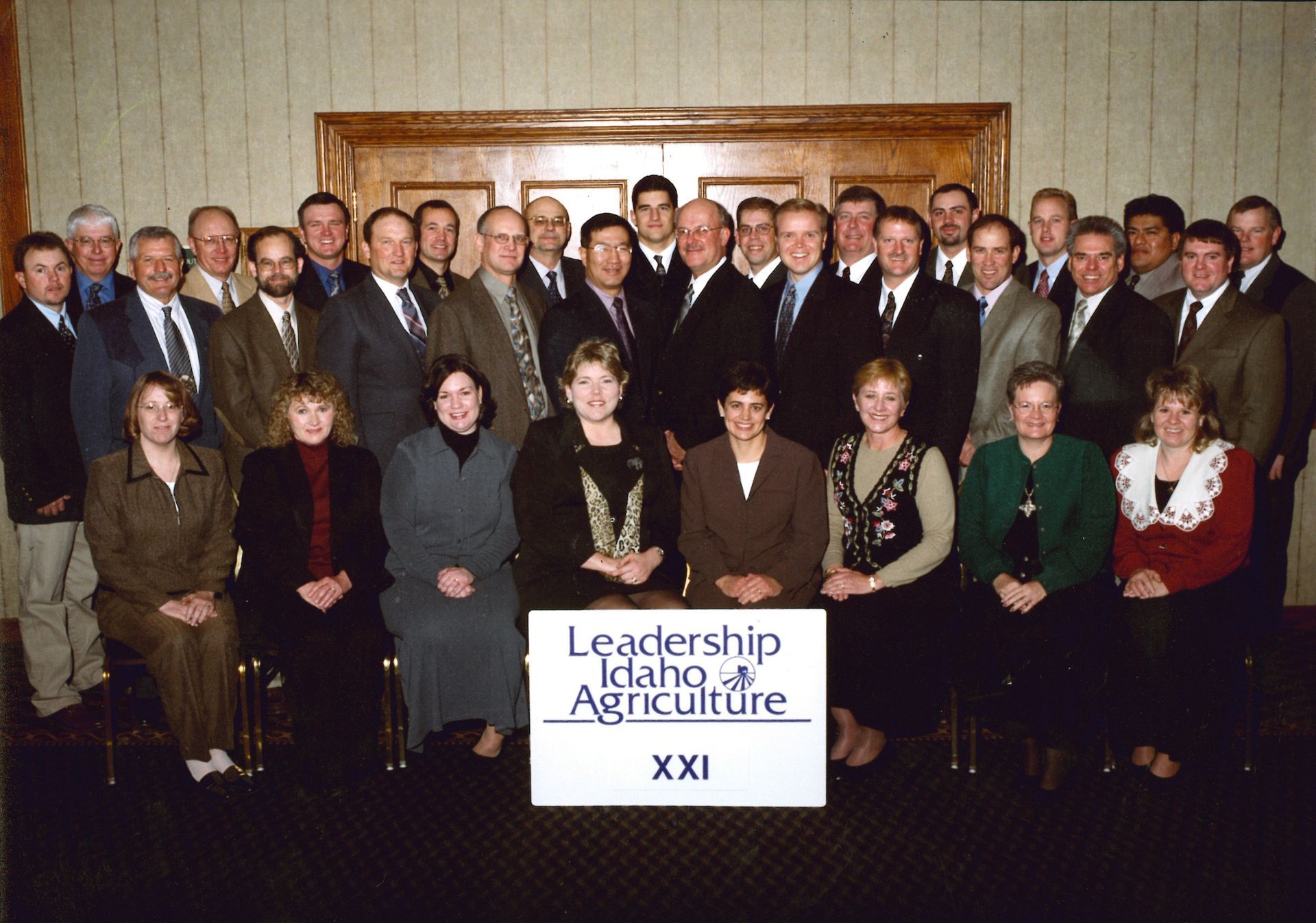 Group portrait of Leadership Idaho Agriculture class XXI members standing and sitting in front of a sign in a room.