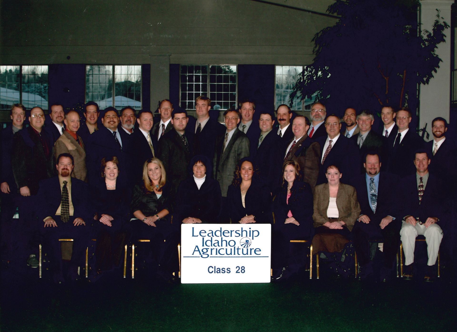 Group portrait of Leadership Idaho Agriculture Class 28 members standing and sitting in front of an event sign indoors.