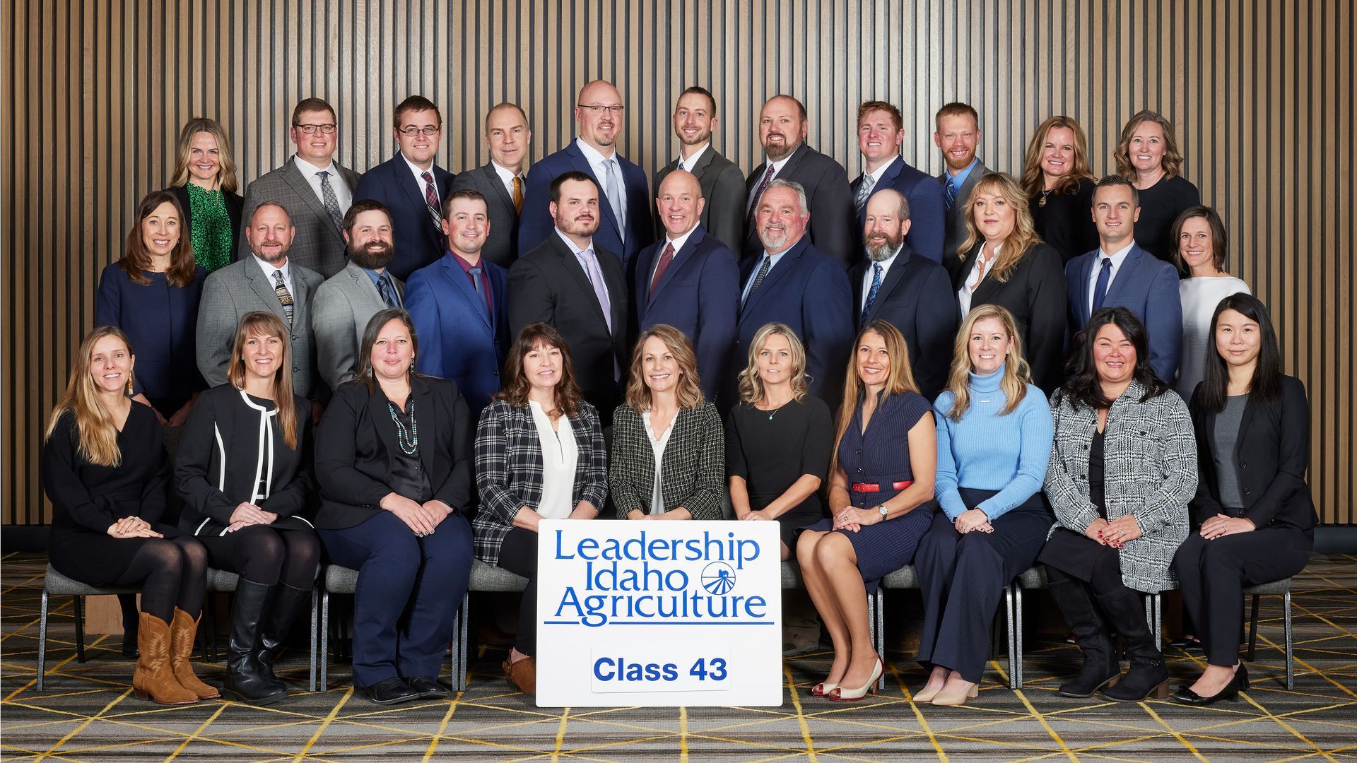 Group portrait of Leadership Idaho Agriculture Class 43 members standing and sitting in front of a wood-paneled wall.