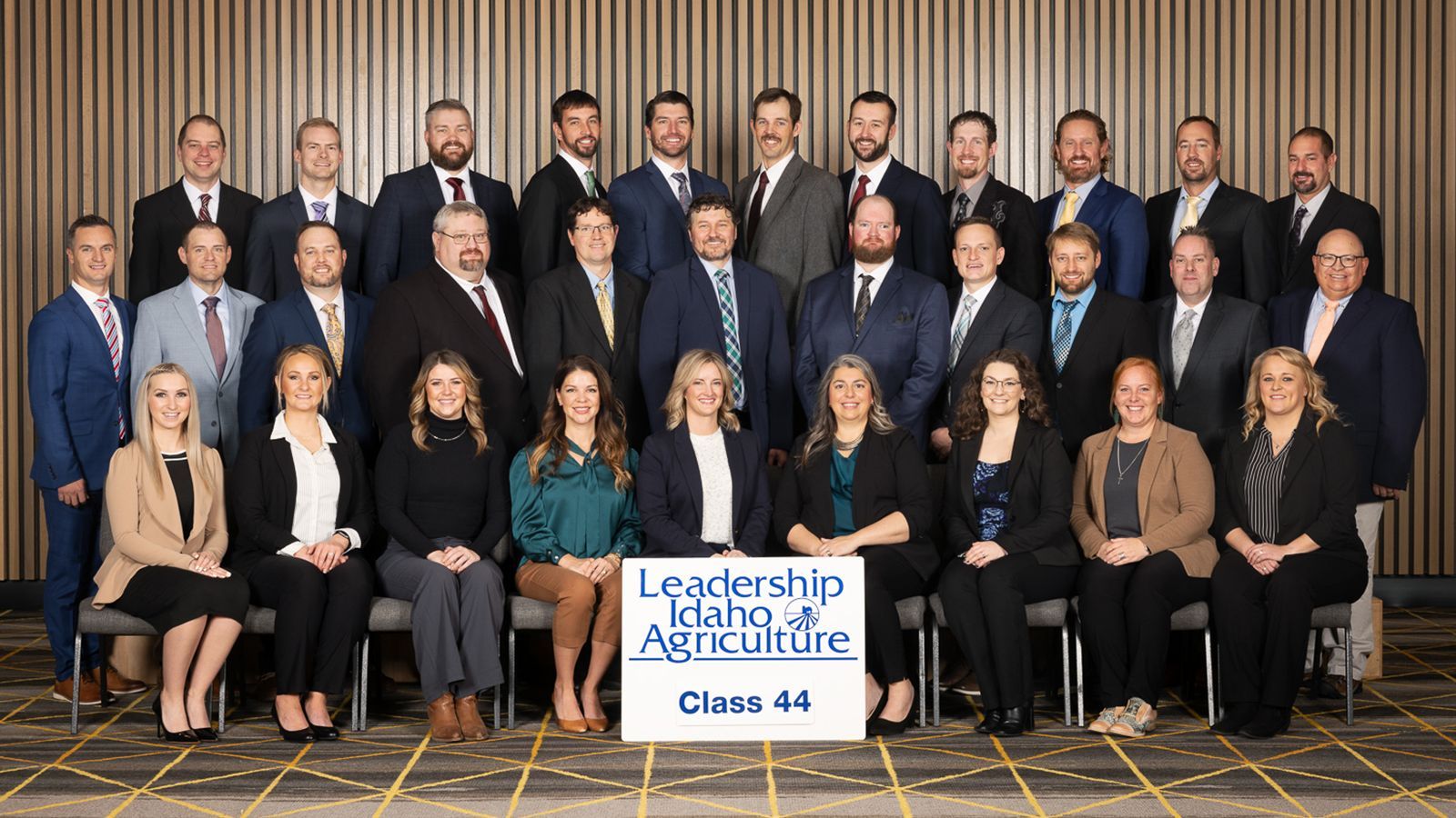A large, formally dressed group poses for a photo in front of a wood-paneled wall with a 