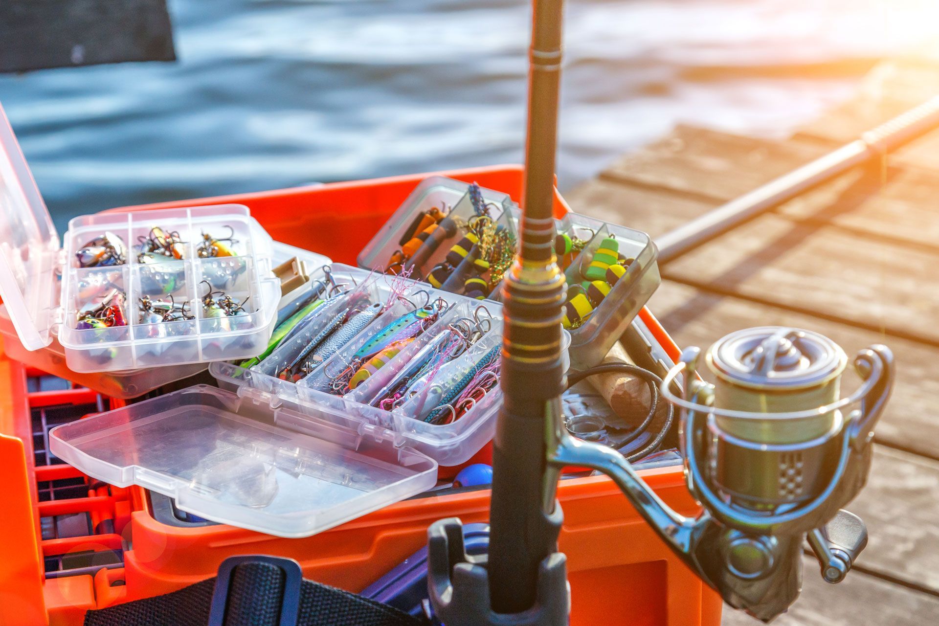 Fishing rod, tackle box with lures, on a wooden dock next to water; sunny day.