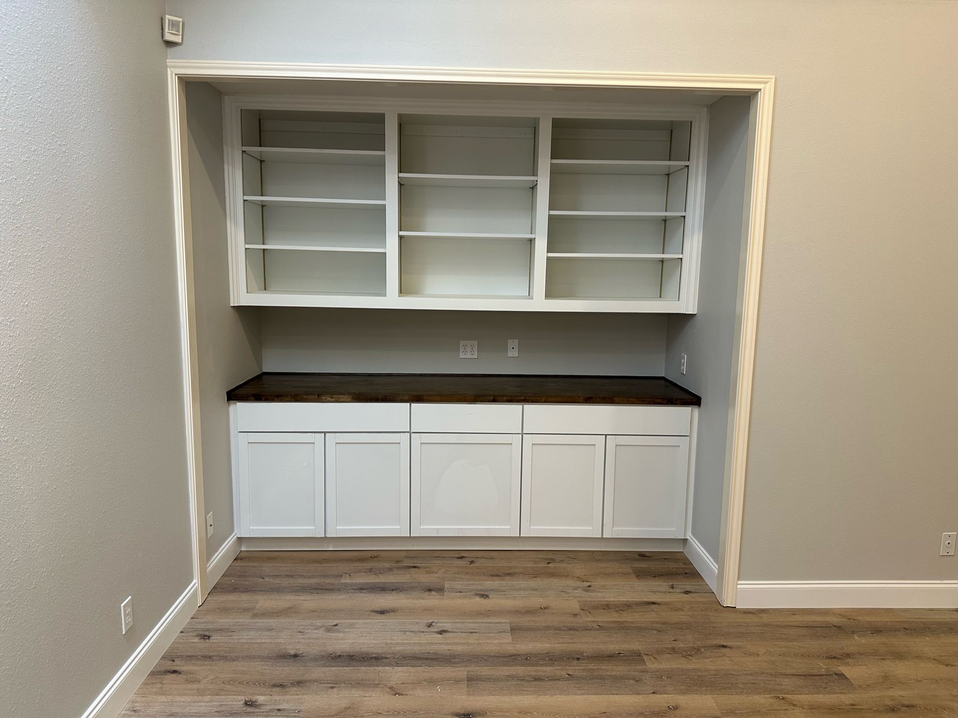 Built-in white bookcase and cabinets with dark countertop, in a room with wood-look flooring and gray walls.