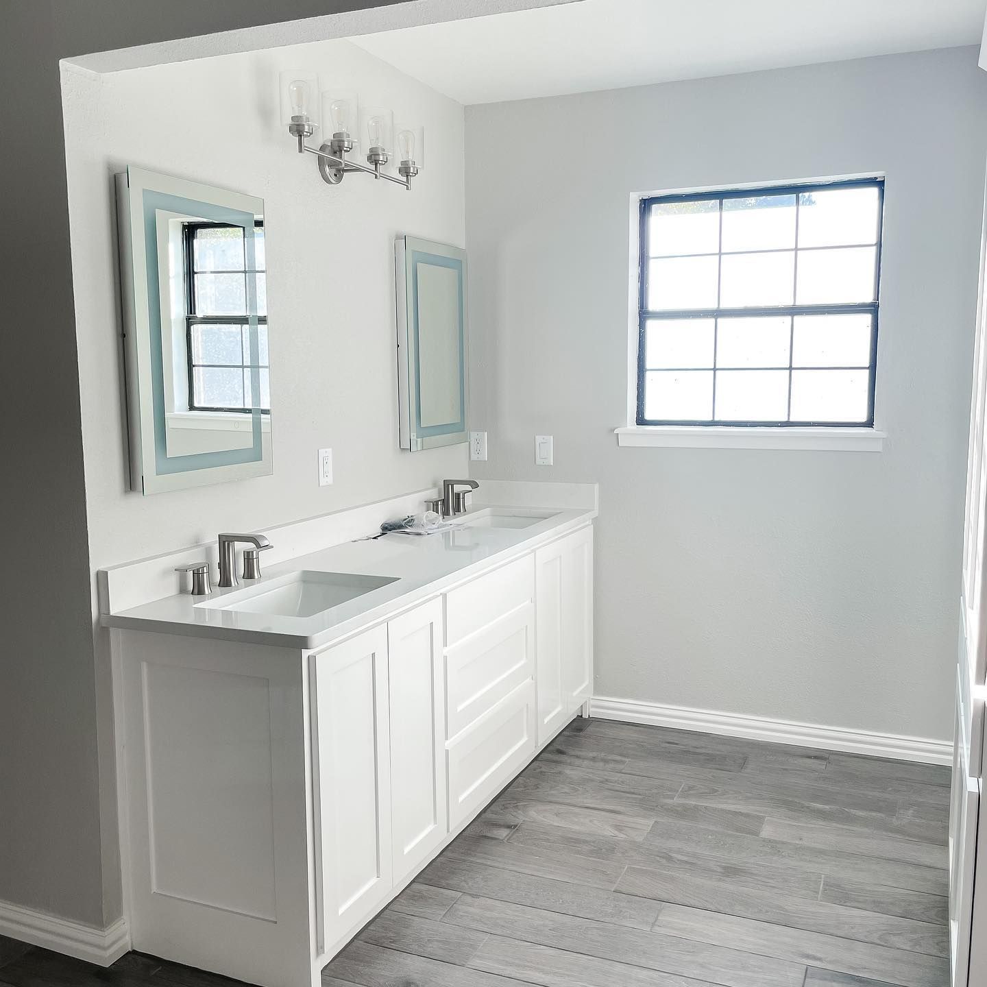 A spacious, well-lit bathroom with a white double vanity, two mirrors, and a window. Gray walls and flooring.