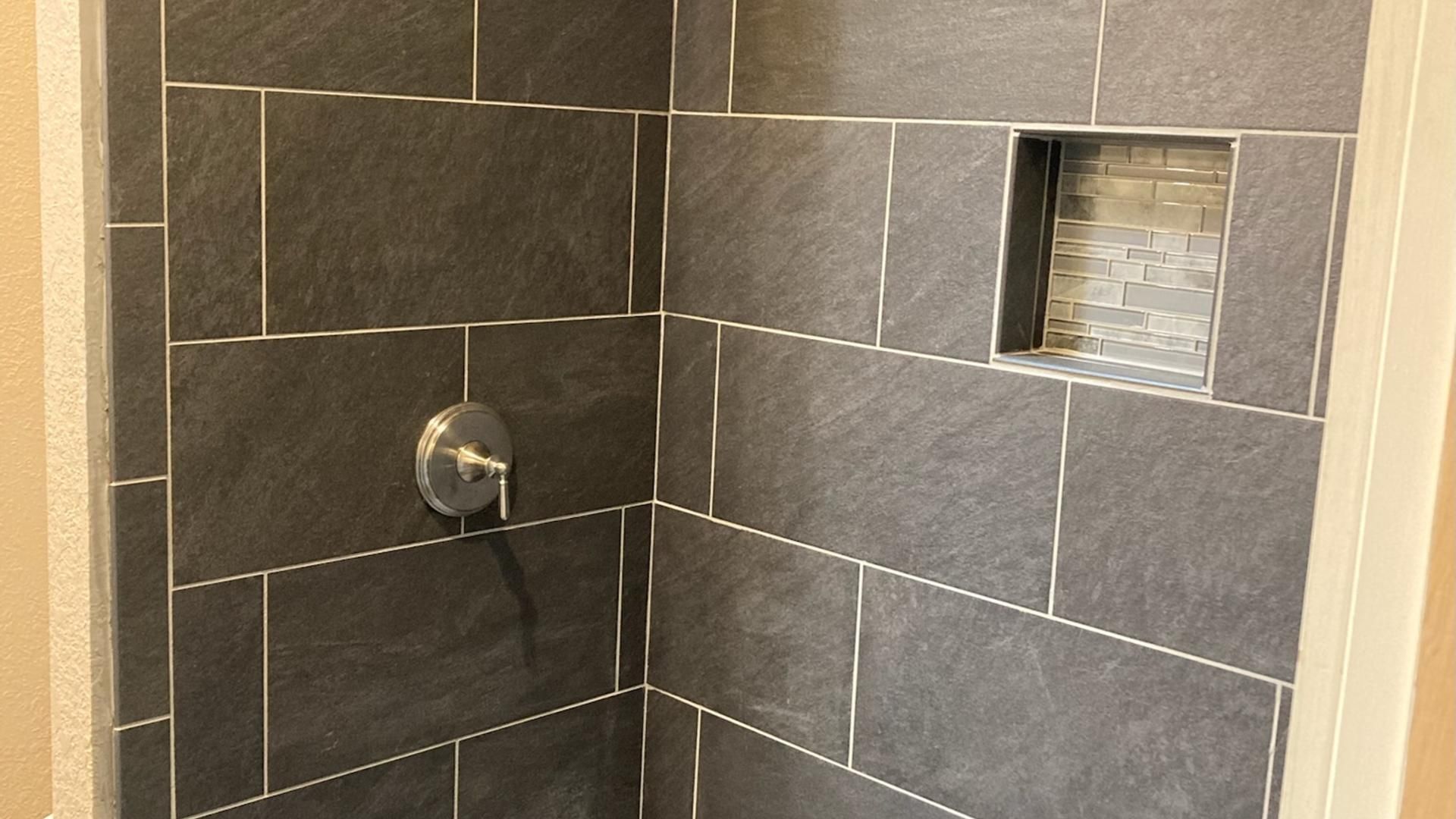 Dark gray tiled shower with built-in shelf; chrome faucet, neutral walls.