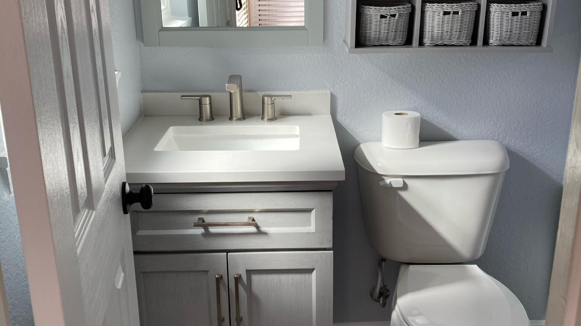Small bathroom with a light gray vanity, white sink, and toilet against a pale blue wall.