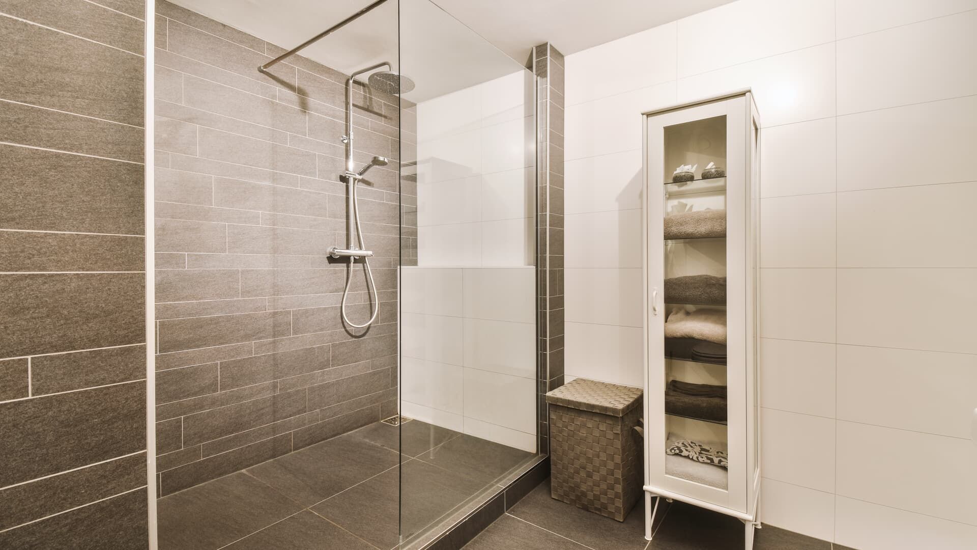 Bathroom with gray tiled shower, glass door, white walls, storage cabinet, and a woven basket.