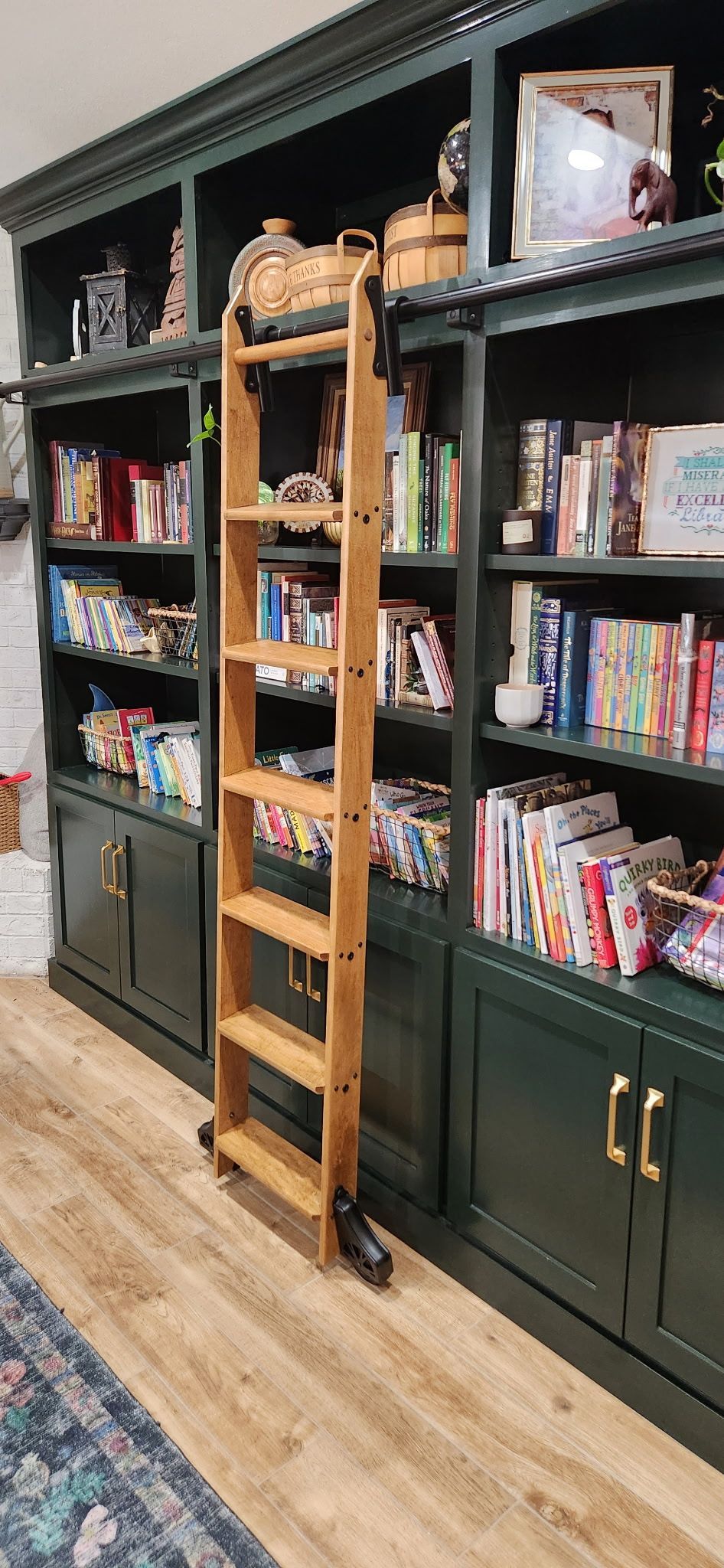 Wooden library ladder on wheels in front of a green built-in bookcase with books and decor.
