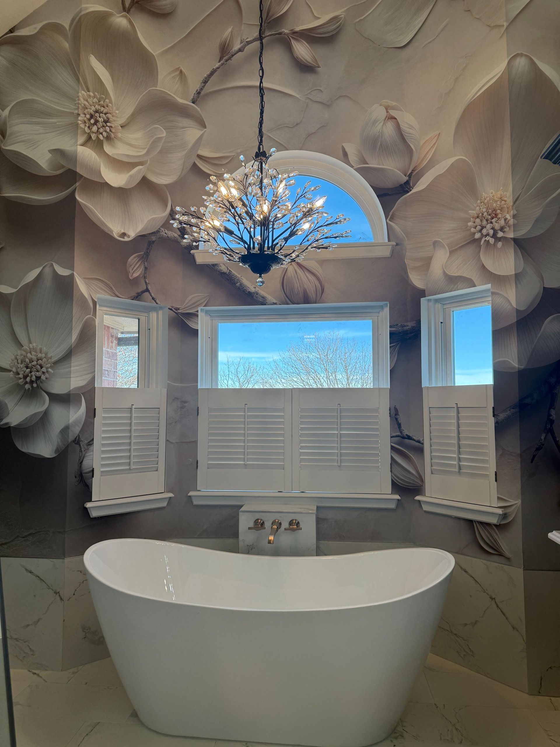 White freestanding tub in a bathroom with floral wall mural and chandelier.