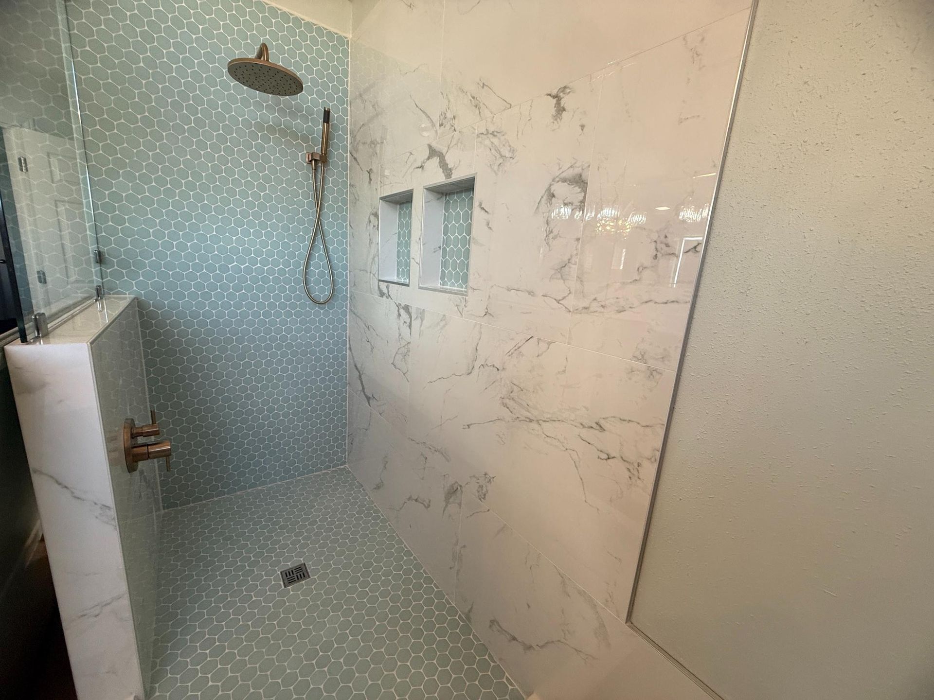 A shower with blue pebble-like tile and marble-patterned walls; bronze fixtures and two built-in shelves.