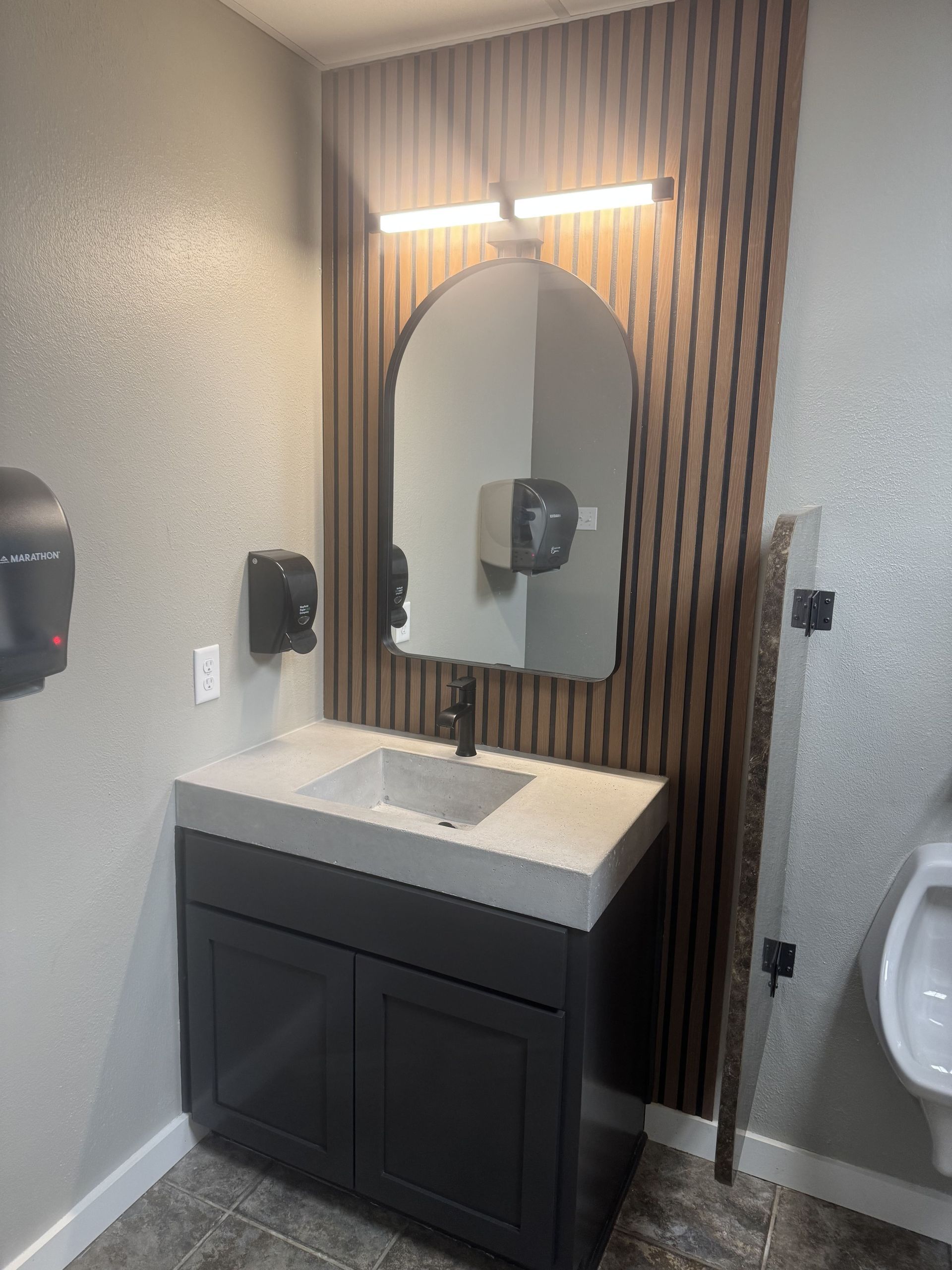 Bathroom vanity with dark gray cabinet, concrete countertop, black faucet, arched mirror, and wood slat accent wall.